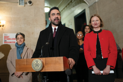 A man speaking at a podium with a city seal, flanked by two women. One woman is smiling and wearing a red jacket, while the other looks on attentively in a gray sweater. In the background, a sign reads 'RENT STABILIZED ORGANIZED'.